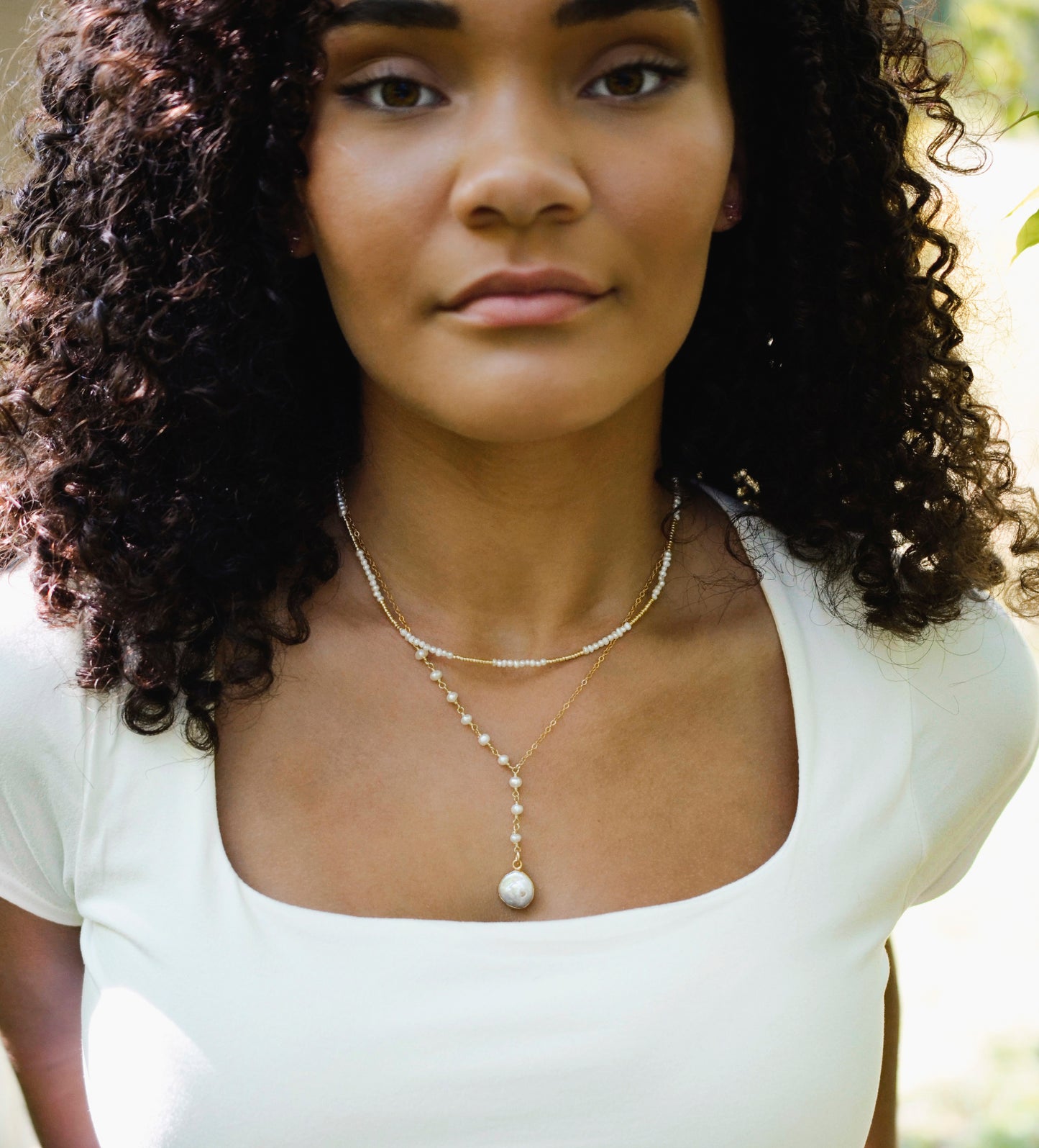 White freshwater pearl necklace with a large coin pearl pendant. Pearls fill one side of the necklace, while the other side is a dainty gold chain. Modeled image. Shown with a beaded pearl choker.