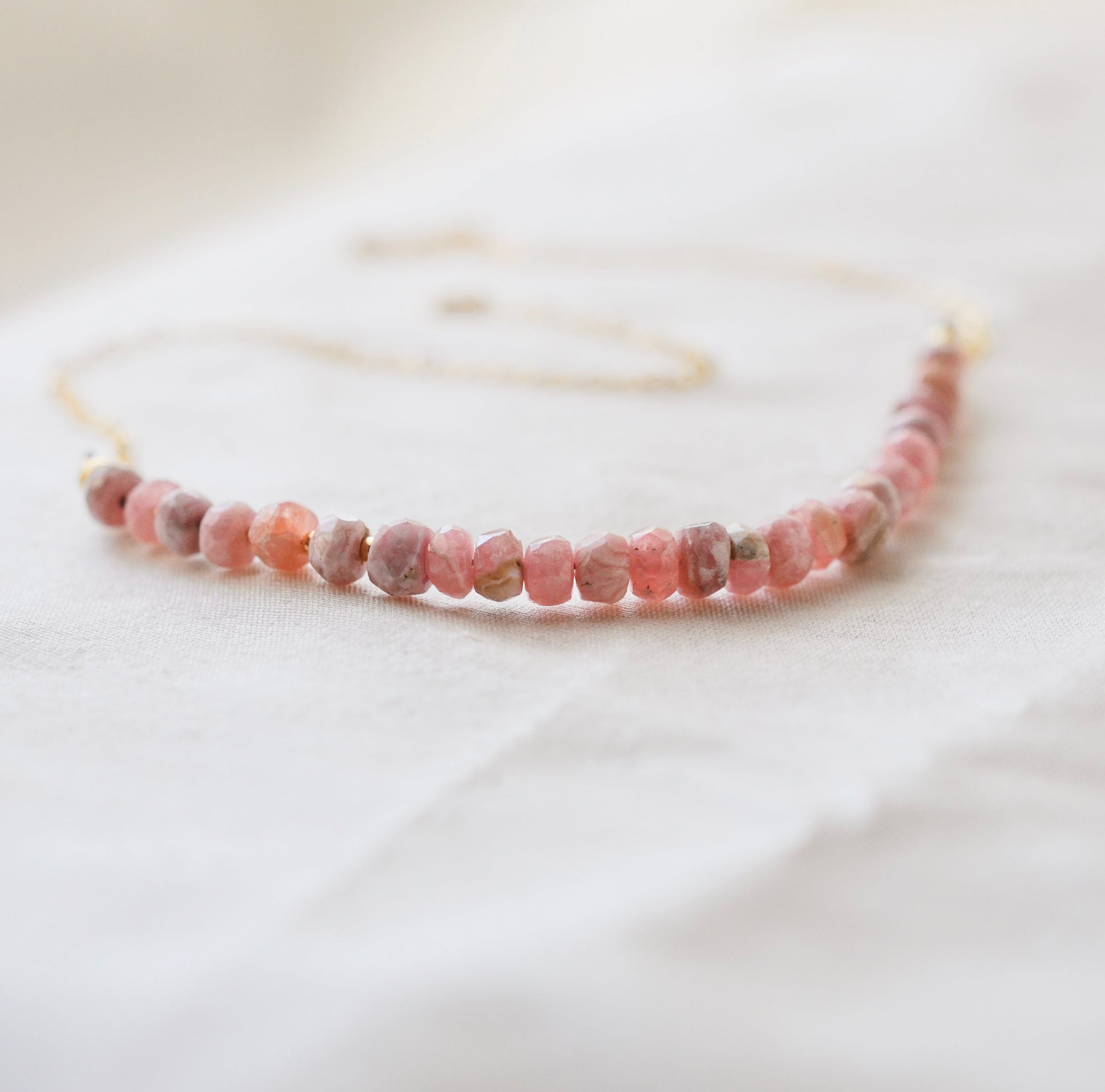 Natural pink Rhodochrosite beaded handmade necklace shown in 14k gold filled. The gemstones vary in shades of pink, peach, white, and gray and are faceted rondelle shape. They span approximately 4 inches across the front of the necklace, then attach to a dainty cable chain. Also available in sterling silver.