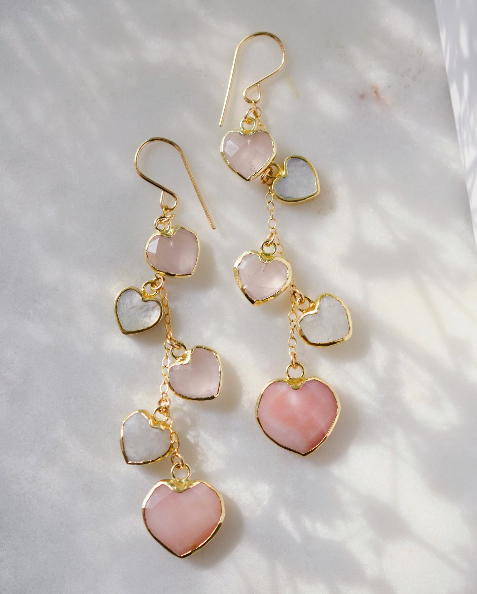 Long pink stone heart earrings in 14k gold filled. There are five heart crystal dangles hanging from a simple chain forming a cluster. The stones include: rose quartz, mother of pearl, pink opal, and rainbow moonstone. They are approximately three inches long.