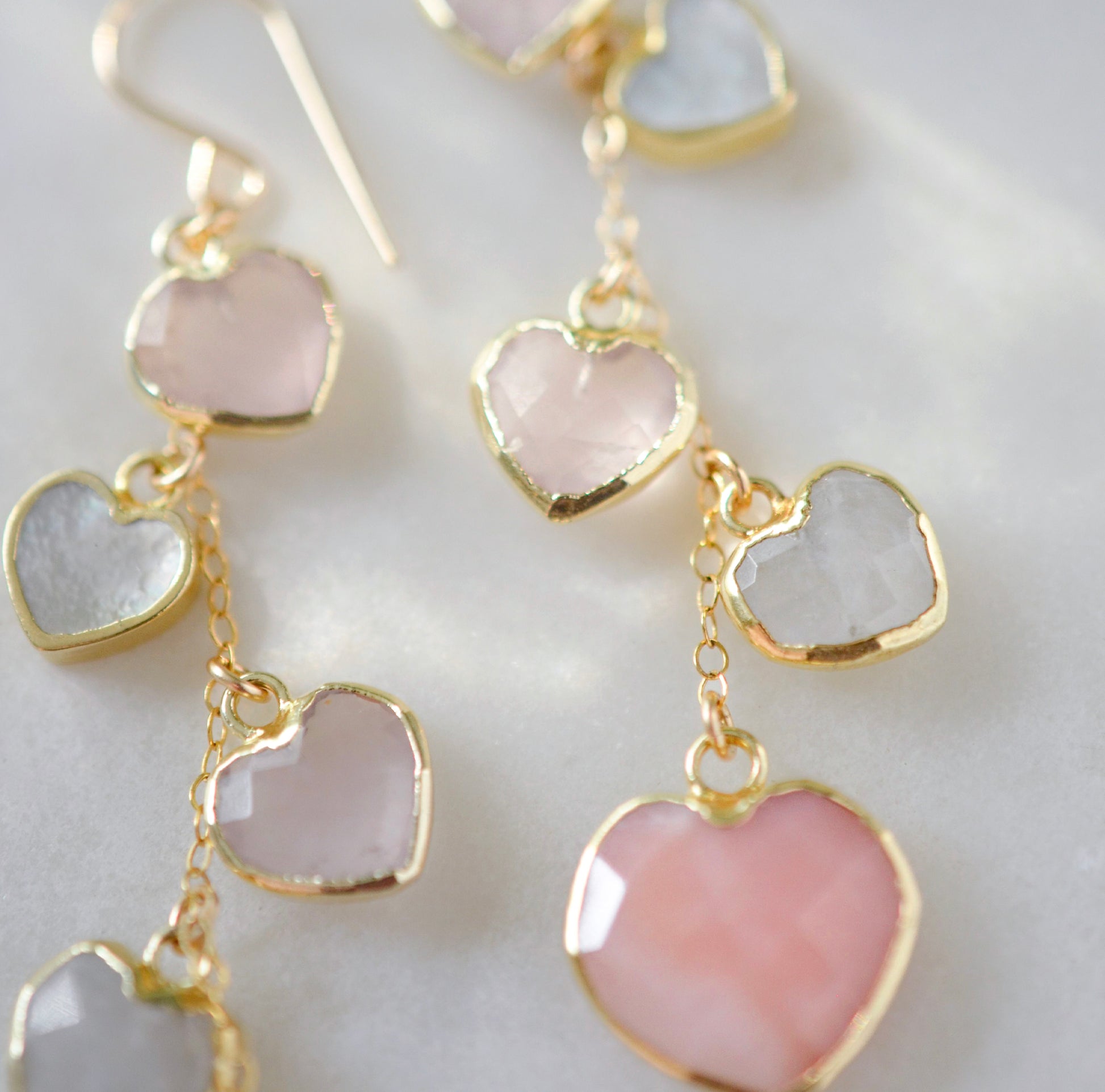 Long pink stone heart earrings in 14k gold filled. There are five heart crystal dangles hanging from a simple chain forming a cluster. The stones include: rose quartz, mother of pearl, pink opal, and rainbow moonstone. They are approximately three inches long.