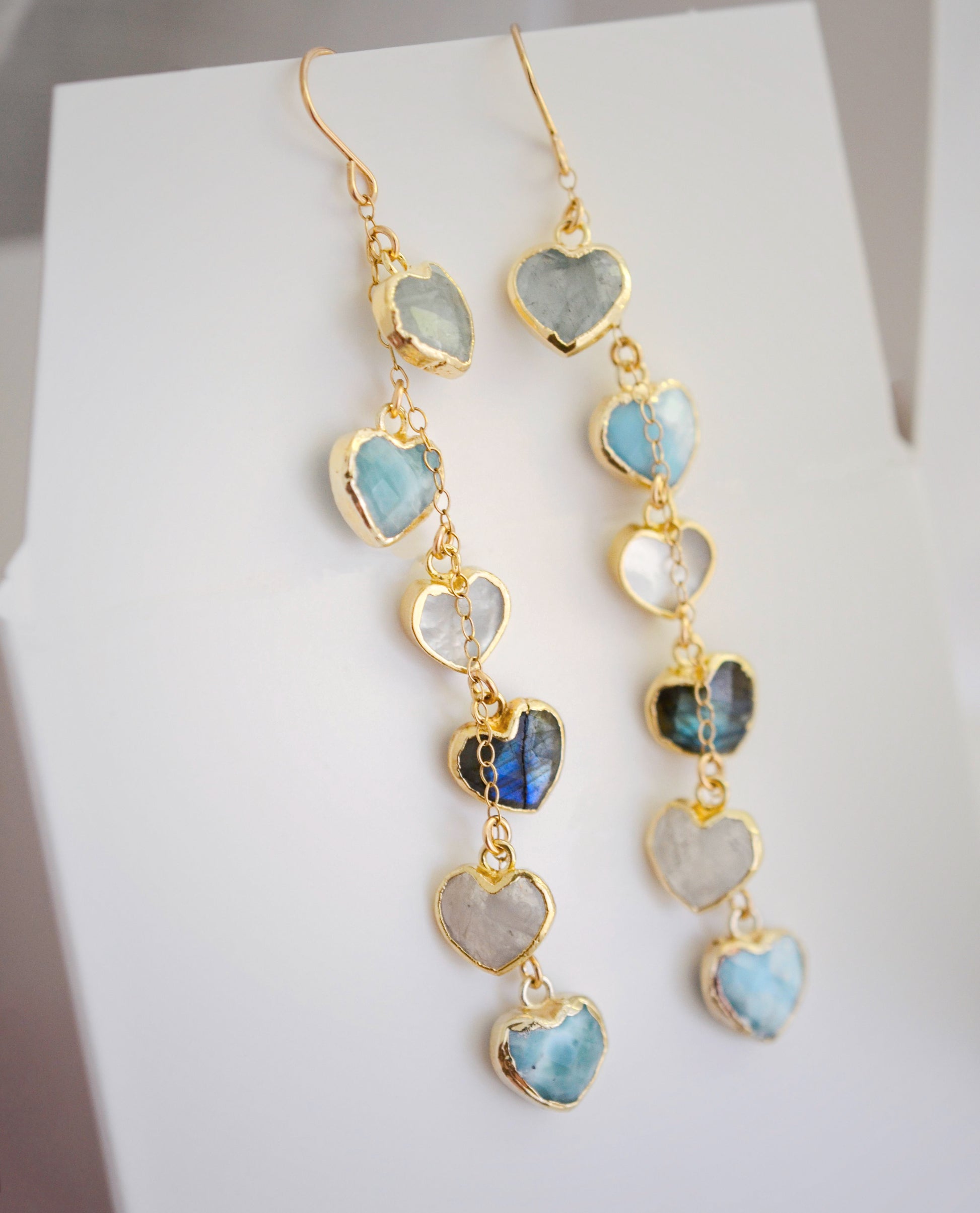 These handmade earrings feature blue and white natural stone hearts bezeled in gold. Each gemstone is a different color and includes: labradorite, larimar, mother of pearl, aquamarine, and rainbow moonstone. Long and light weight they make the perfect romantic gift. They are 3 inches long.
