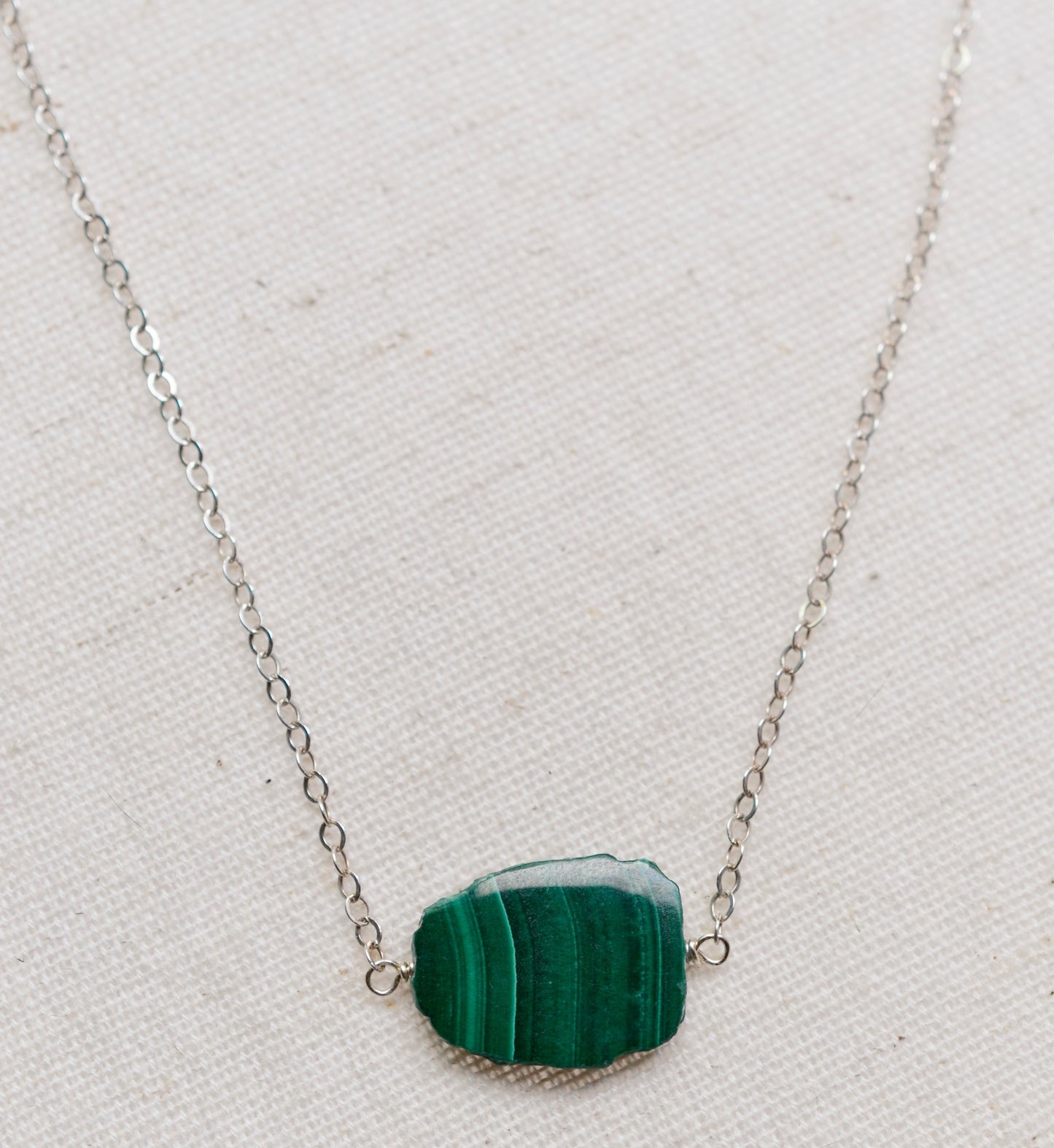 Natural green Malachite smooth polished slice with raw edges. Each stone is an irregular oval shape and has a variety of banding. Shown on a sterling silver chain.