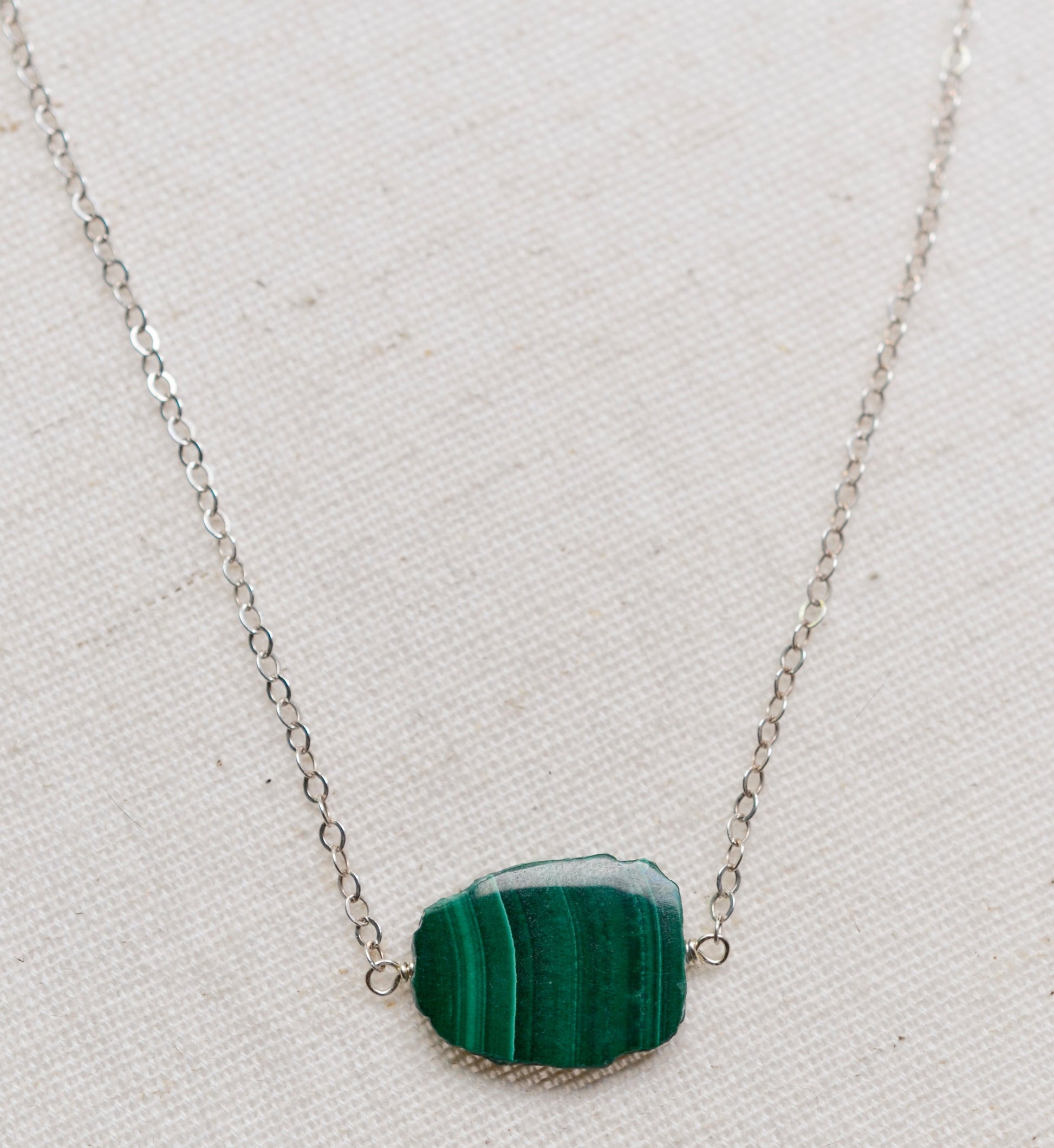 Natural green Malachite smooth polished slice with raw edges. Each stone is an irregular oval shape and has a variety of banding. Shown on a sterling silver chain.