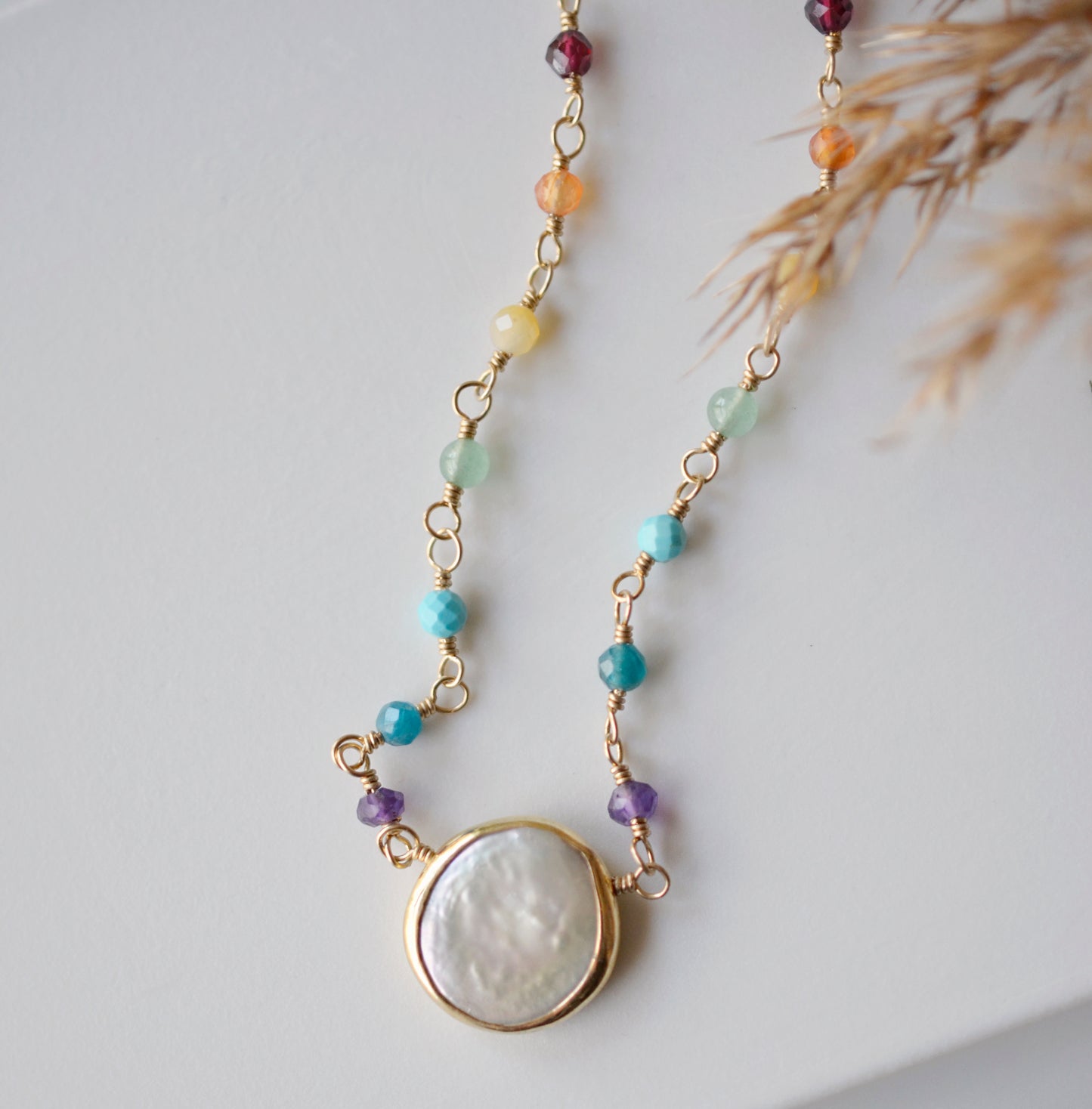 Freshwater pearl and rainbow chakra gemstone necklace. A white pearl, bezeled in gold or sterling silver sits at the center of the necklace below a handmade chain of rainbow stones. The gold style is shown.