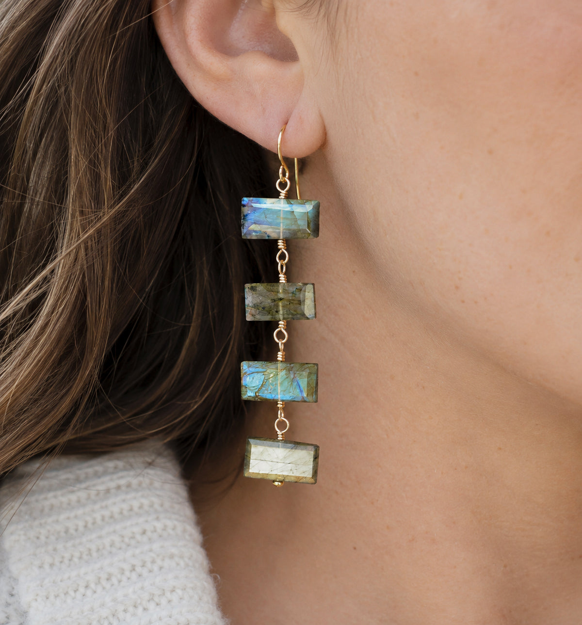 A close-up of a woman wearing a long dangle earring made of four rectangular labradorite stones in blue-green tones with 14k gold filled hooks and links.