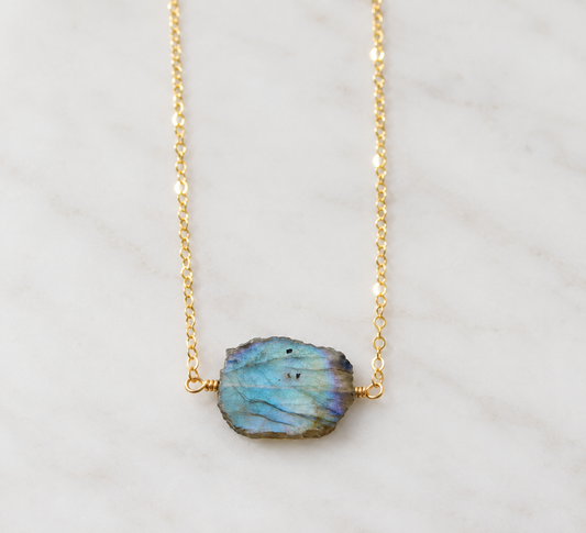 A 14k gold filled chain necklace featuring a raw, irregular-shaped labradorite stone pendant with shimmering blue and green iridescence. The edges are rough, but the crystal face is smooth polished on both sides. Dainty handmade gemstone jewelry by Gemnia.