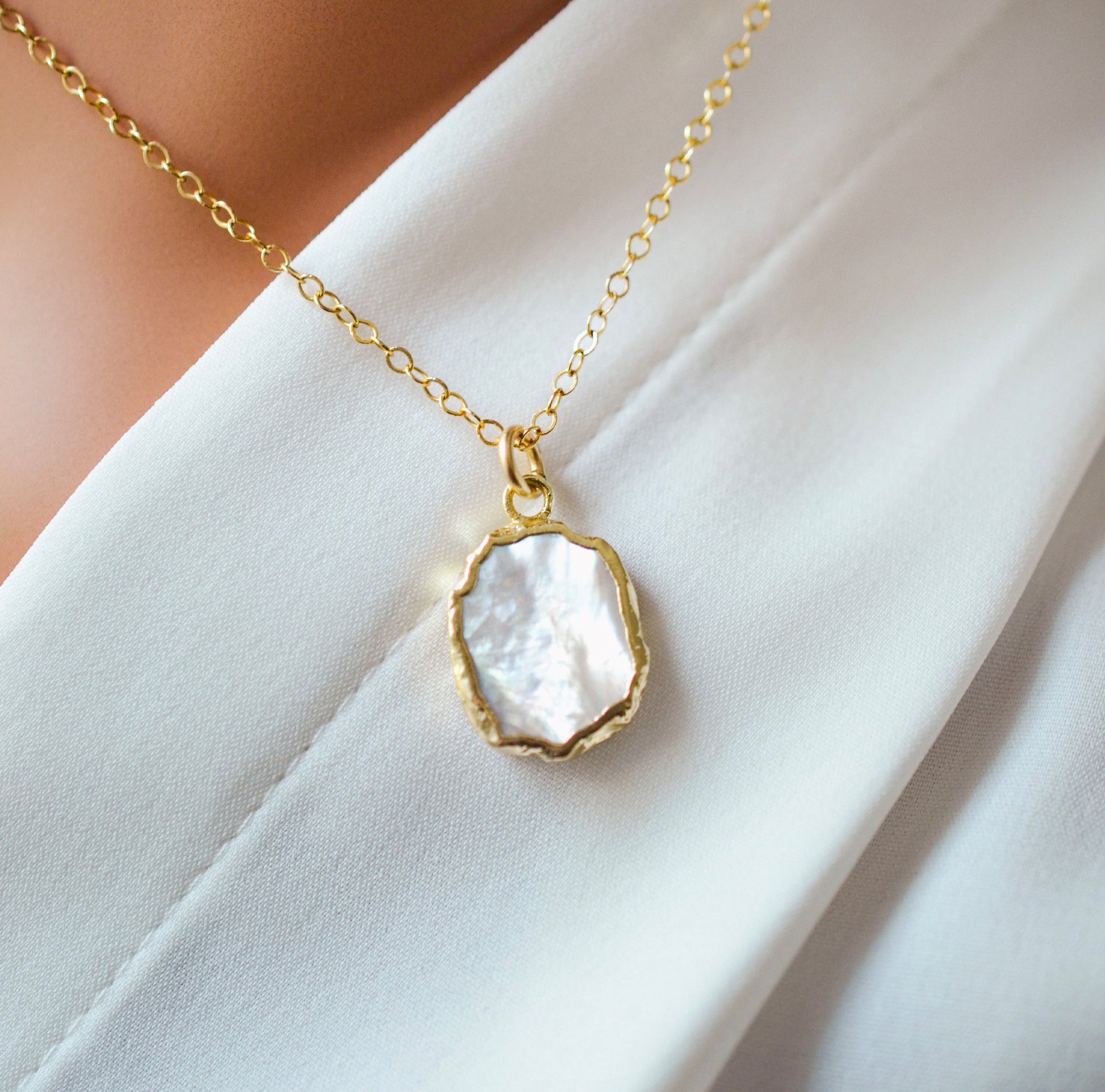 A smooth polished natural, white mother of pearl slice has been set in a 24k gold electroplated bezel and suspended from a 14k gold filled chain. The shell shimmers in shades of white, silver, and pink. It is shown on white fabric.