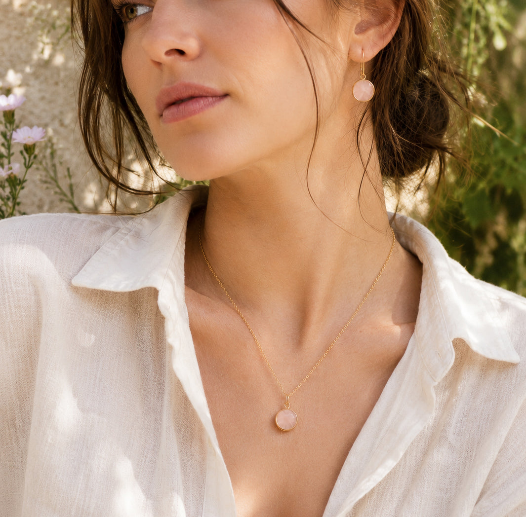 A woman modeling a natural rose quartz earrings and pendant in 14k gold filled. The gemstones are a pale, translucent pink and also available in sterling silver. 