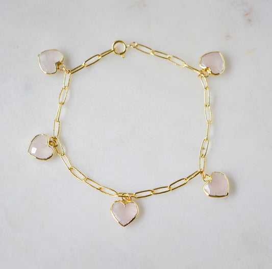 A gold paperclip chain bracelet with heart-shaped pink rose quartz gemstones on a light gray background. 