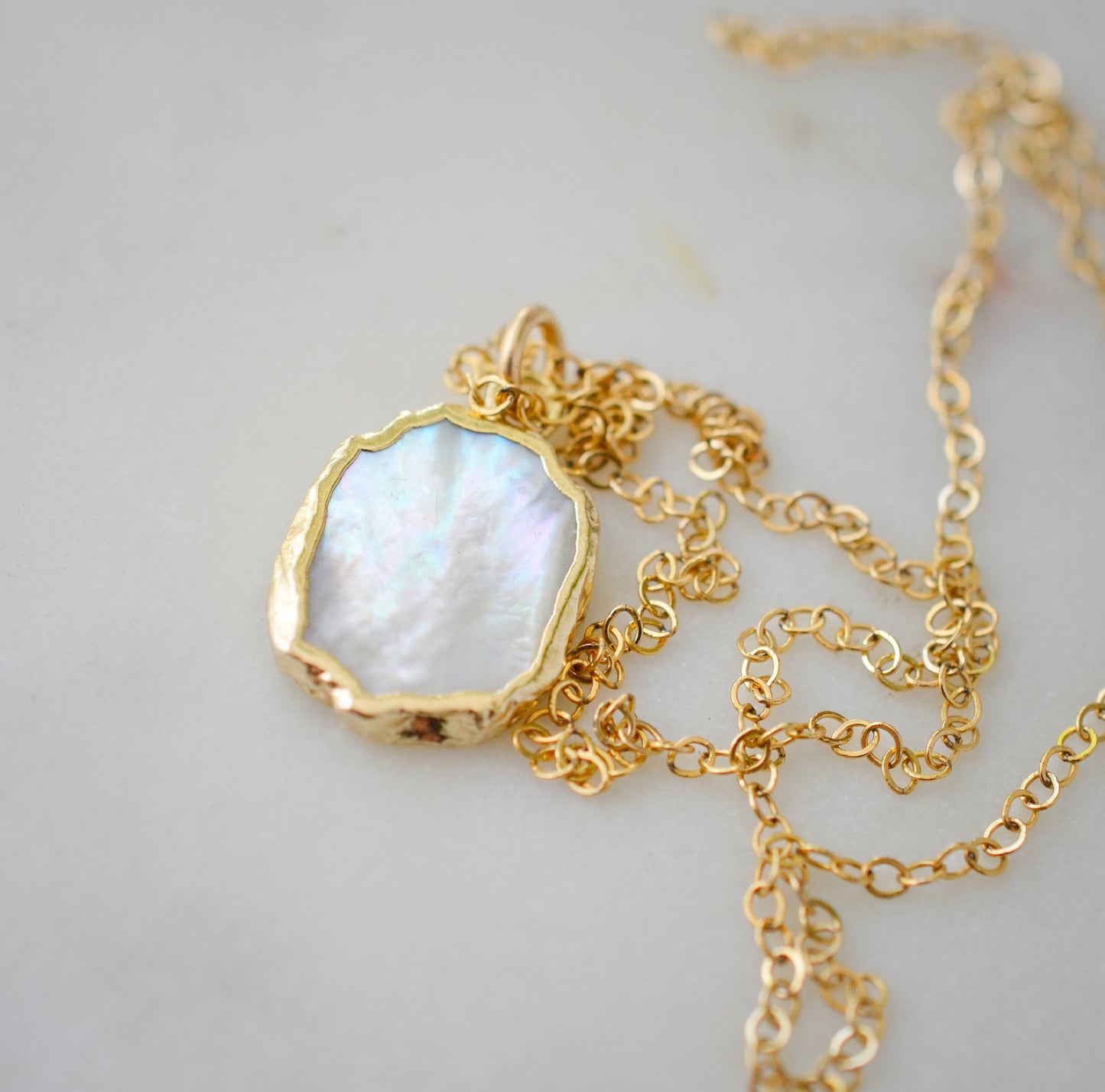 Close up image. A smooth polished natural, white mother of pearl slice has been set in a 24k gold electroplated bezel and suspended from a 14k gold filled chain. The shell shimmers in shades of white, silver, and pink.