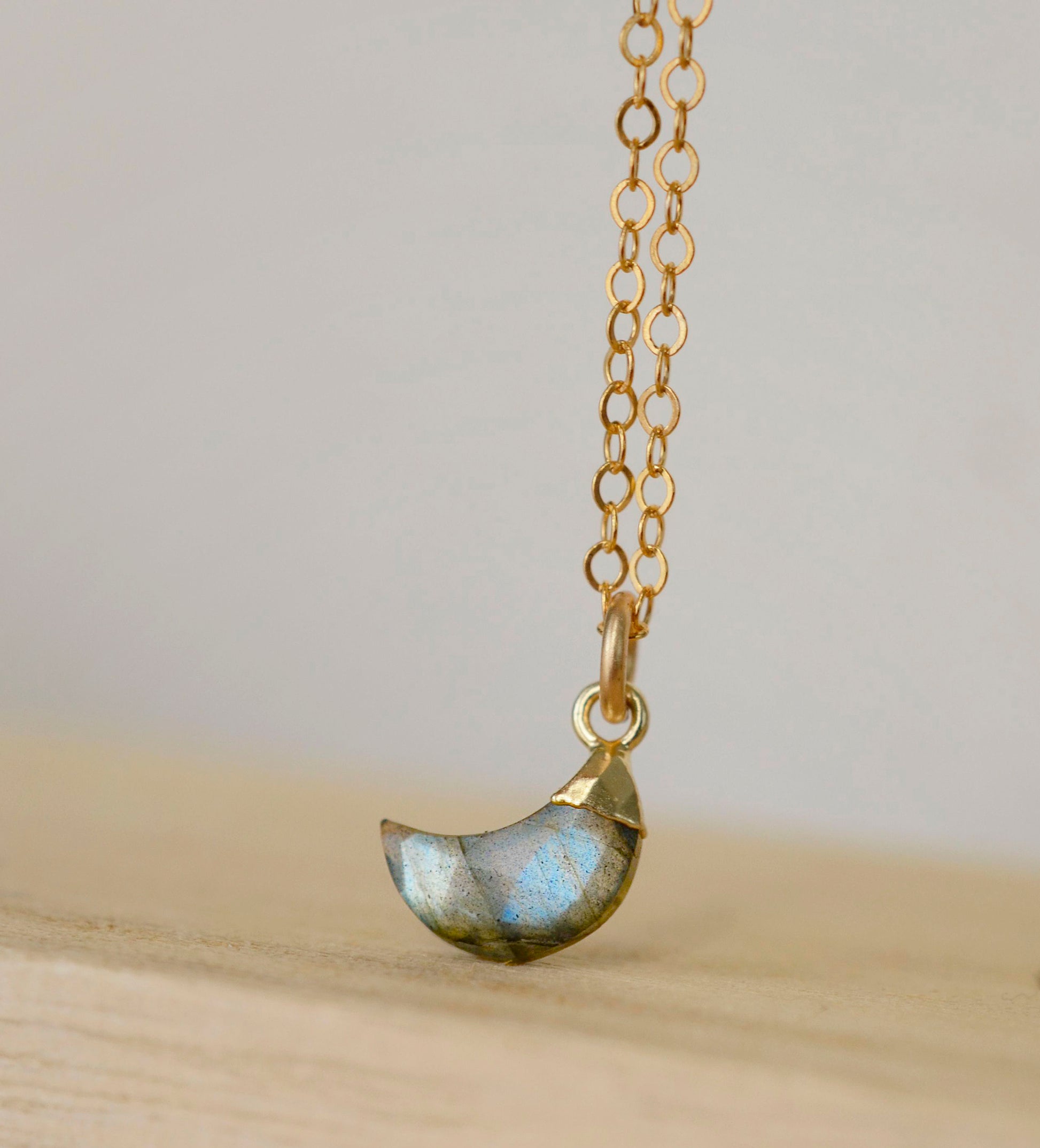 A close up image of a natural labradorite gemstone shaped in a crescent moon and set on a 14k gold filled chain. 