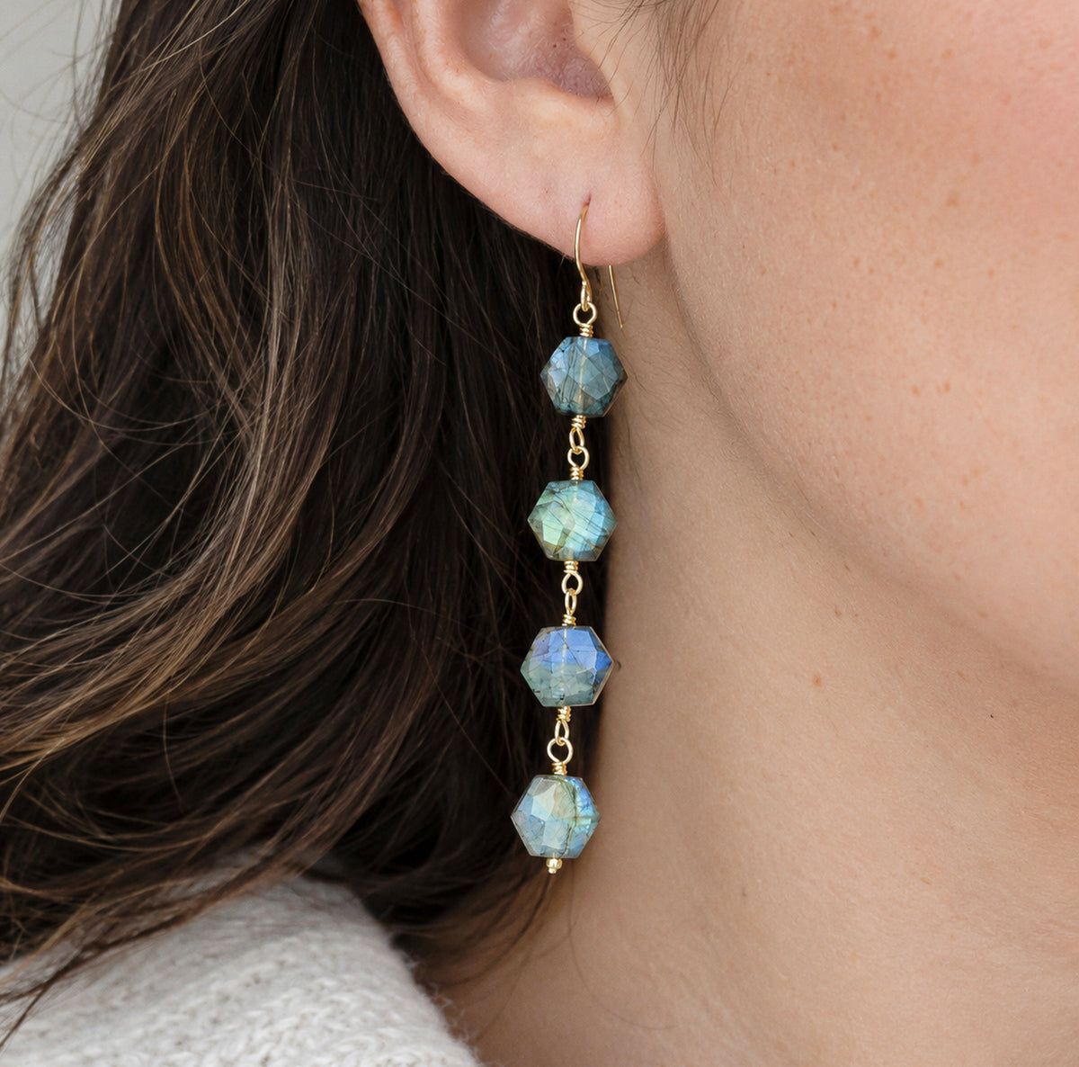 A closeup of a woman wearing long labradorite dangle earrings with hexagon shaped gemstones in 14k gold filled. Handmade in Connecticut by Gemnia.