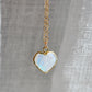 Heart-shaped rainbow moonstone pendant on a 14k gold filled chain against a gray fabric background. This dainty romantic necklace is handmade by Gemnia Jewelry in Connecticut.
