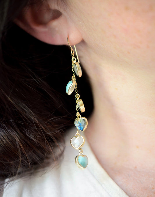 Modeled image. These handmade earrings feature blue and white natural stone hearts bezeled in gold. Each gemstone is a different color and includes: labradorite, larimar, mother of pearl, aquamarine, and rainbow moonstone. Long and light weight they make the perfect romantic gift. They are 3 inches long.