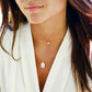 A woman wearing a white mother of pearl oval pendant on a 14k gold filled chain layered with a dainty, minimalist freshwater pearl necklace.