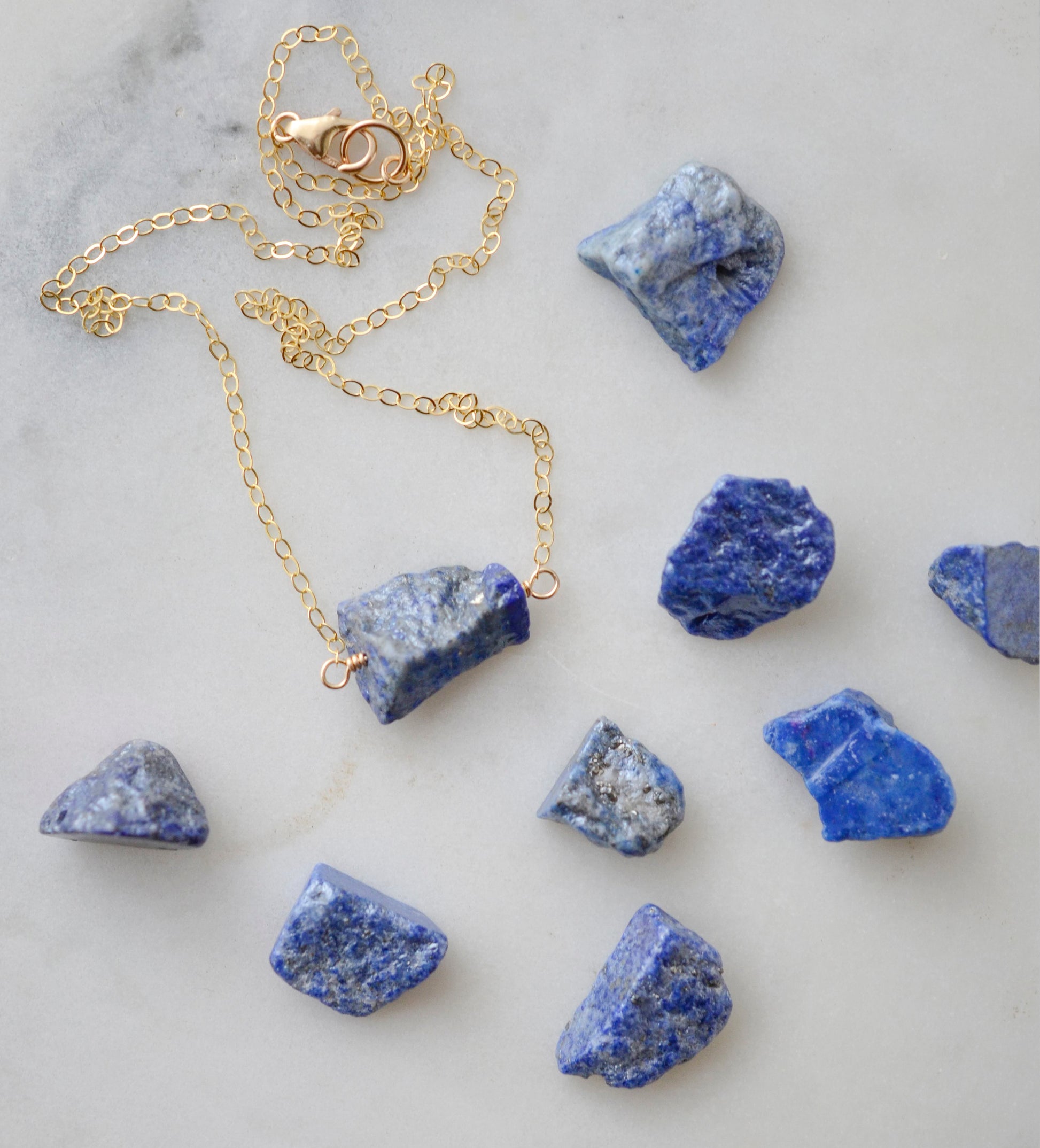 Raw blue lapis lazuli stone suspended from a gold chain. Other available stones shown.