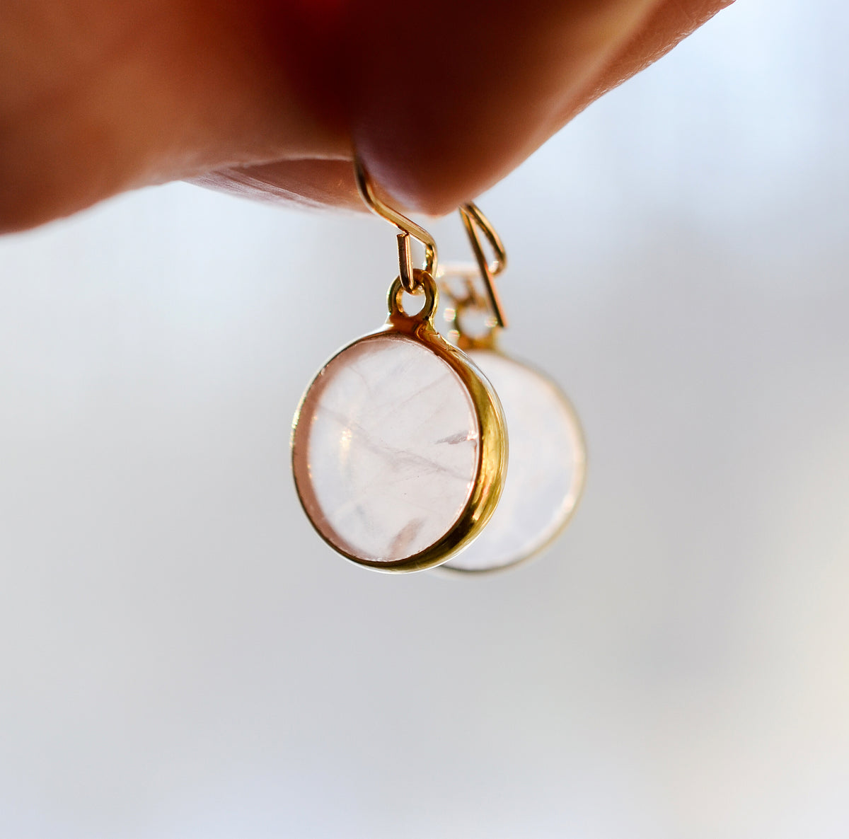 A close-up of a hand holding a pair of gold dangle earrings with round, natural, pale pink rose quartz gemstones.