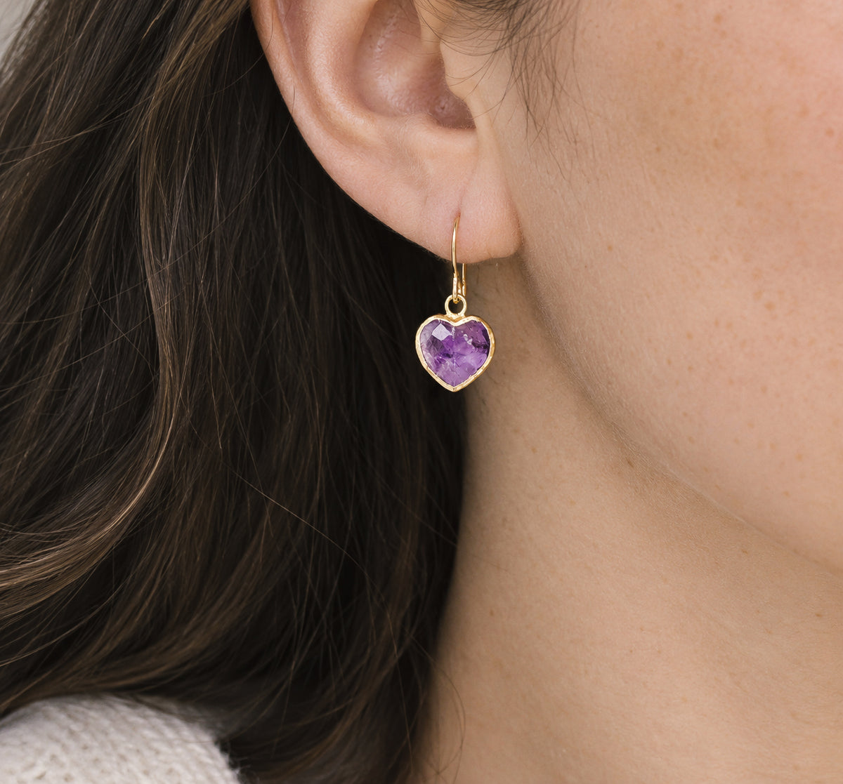 A woman wearing natural purple amethyst heart shaped dangle earrings in gold. The bezel is 18k gold electroplate and is organic in shape. The earwires are french hooks. Handmade jewelry by Gemnia.