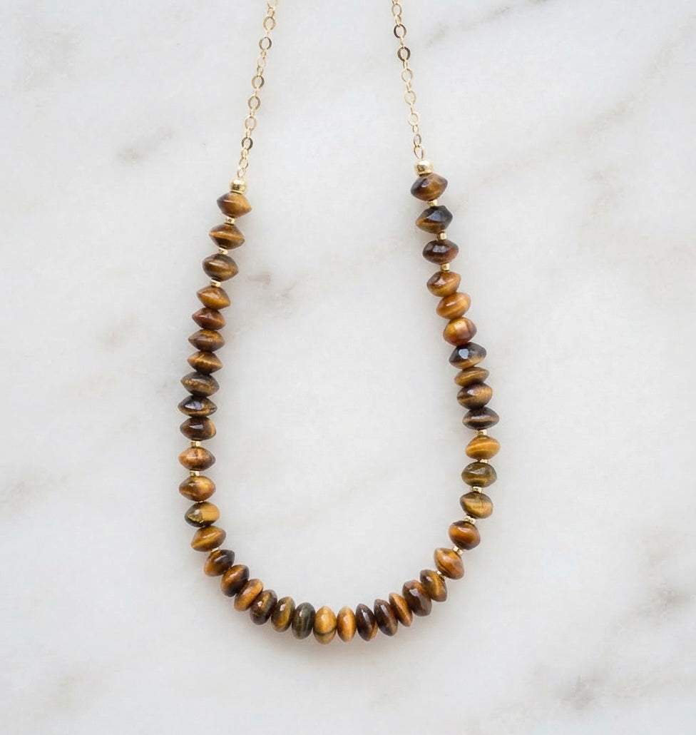A beaded necklace made of polished tiger's eye beads on a delicate gold chain, displayed on a white marble surface. Handmade jewelry in USA by Gemnia.