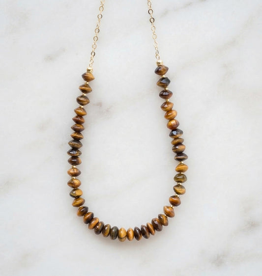 A beaded necklace made of polished tiger's eye beads on a delicate gold chain, displayed on a white marble surface. Handmade jewelry in USA by Gemnia.