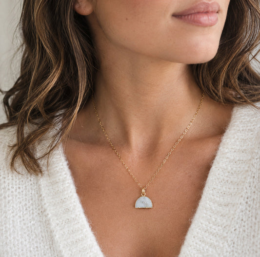 A close-up of a woman wearing a delicate gold chain necklace with a small, semi-circular white pendant featuring a natural sparkling white quartz druzy. Handmade in Connecticut by Gemnia.