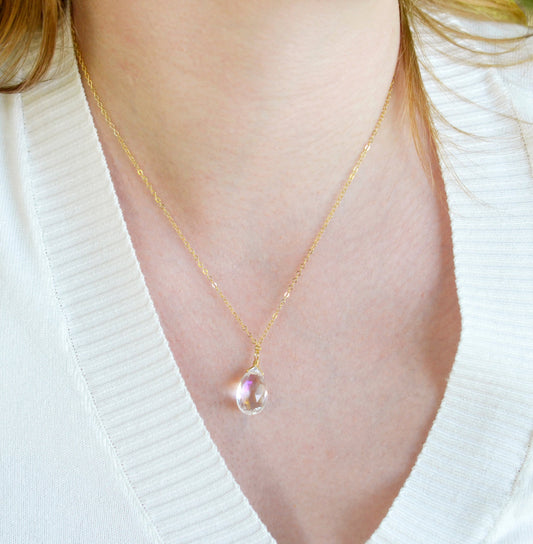 A woman wearing a clear, faceted mystic topaz teardrop pendant with shimmering rainbow colors within the gemstone. This dainty necklace is set on a simple 14k gold filled chain. Handmade jewelry in Connecticut by Gemnia.