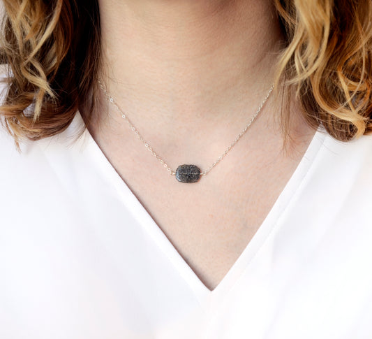 A glittering black sunstone smooth polished slice with raw edges set onto a sterling silver chain. Modeled image. Stone shimmers a mixture of black and silver.