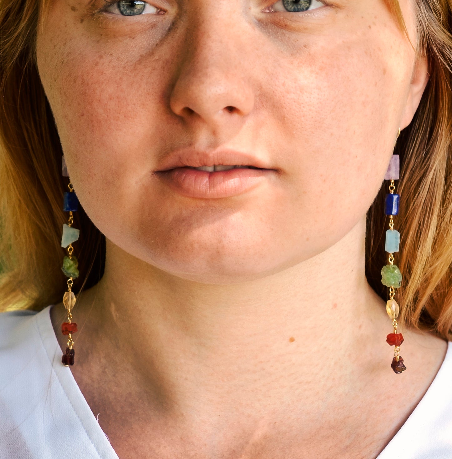 Long gemstone earrings made with a rainbow of raw gemstones. Colors include: purple, blue, aqua, green, yellow, orange, and red. Modeled image. 